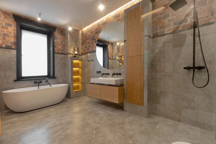 Spacious wet room with exposed brick accent wall, freestanding tub, wall-mounted wood vanity with vessel sink, illuminated recessed shelving, and open glass shower area.