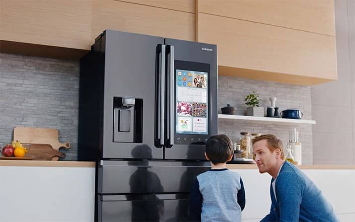 A father and son kneel in front of a smart refrigerator with a screen on the door.