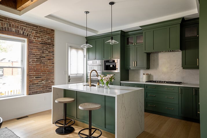 Exposed brick wall complemented by dark leafy green cabinets and white countertops.