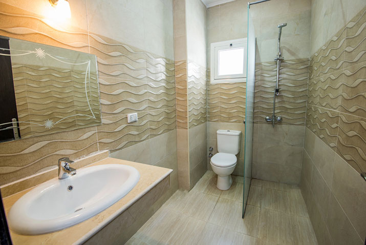 Compact wet room in an apartment with wave-textured beige wall tile, a glass shower partition, wall-mounted sink, and seamless tile flooring.