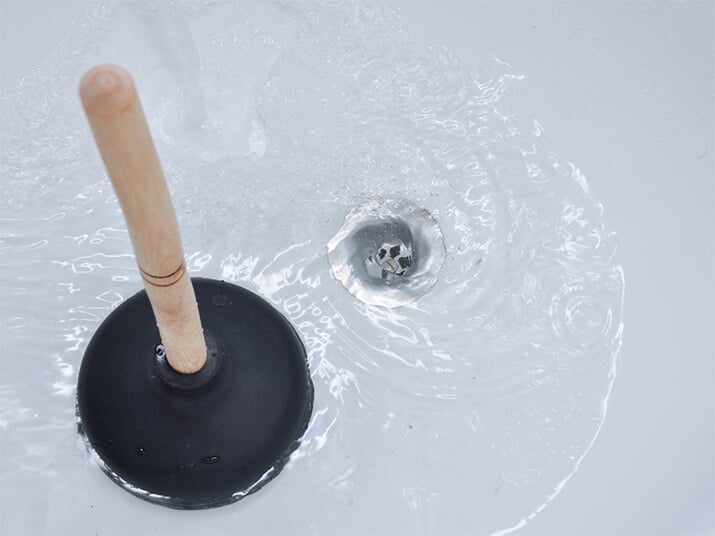A plunger sits by a drain around which water is swirling.