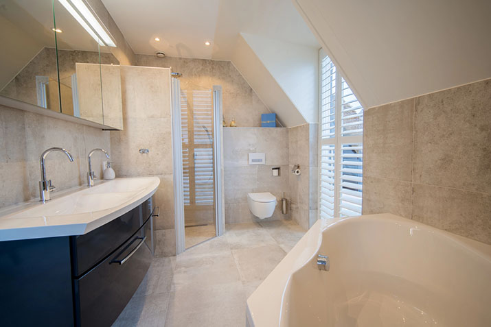 Wet room bathroom under a sloped ceiling with a dark double vanity, wall-mounted toilet, glass-enclosed shower, soaking tub, and large stone tiles throughout.
