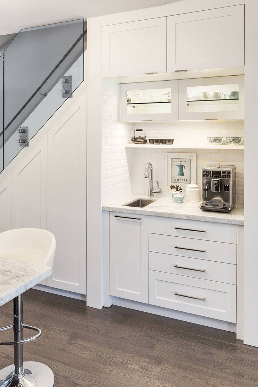 White coffee station built under the stairs with subway tile and a sink.