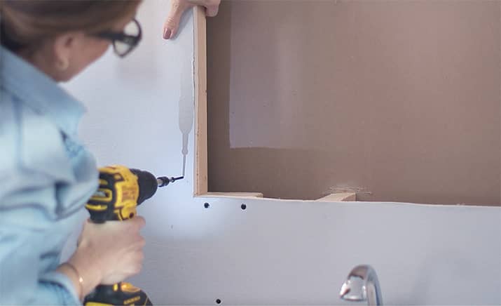 A woman adds a block of wood to the drywall of a recessed medicine cabinet.
