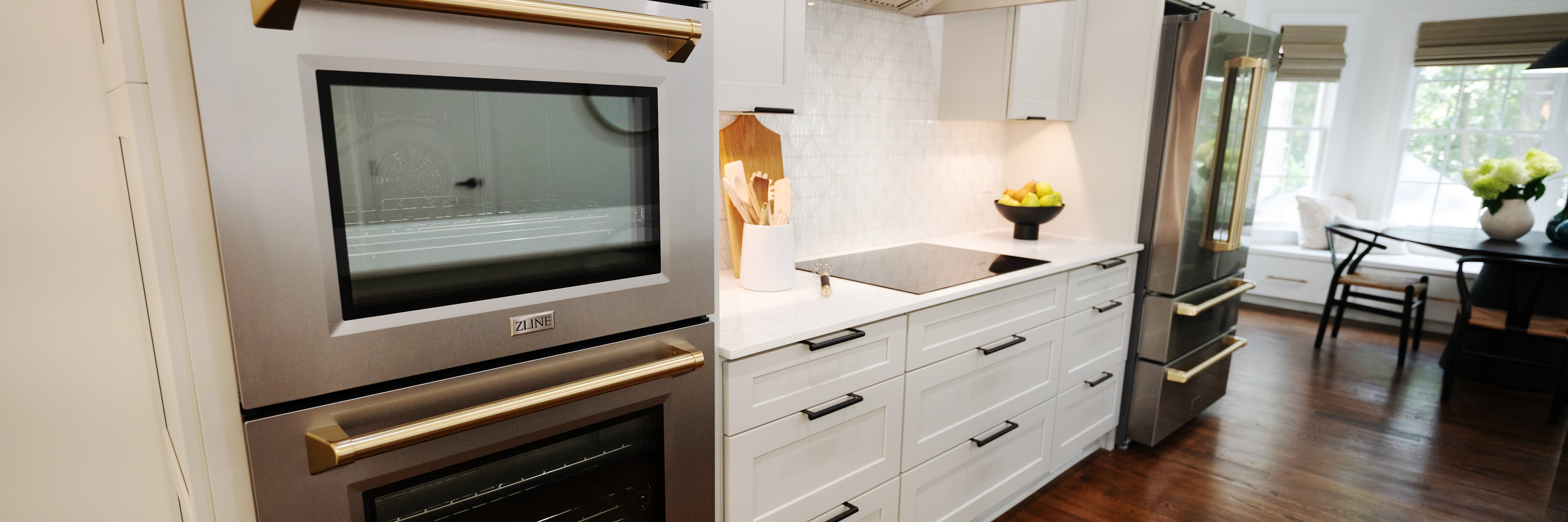 A Shaker Light Gray kitchen with brass-handled wall ovens, black drawer pulls, and a sunny breakfast nook, renovated on HGTV