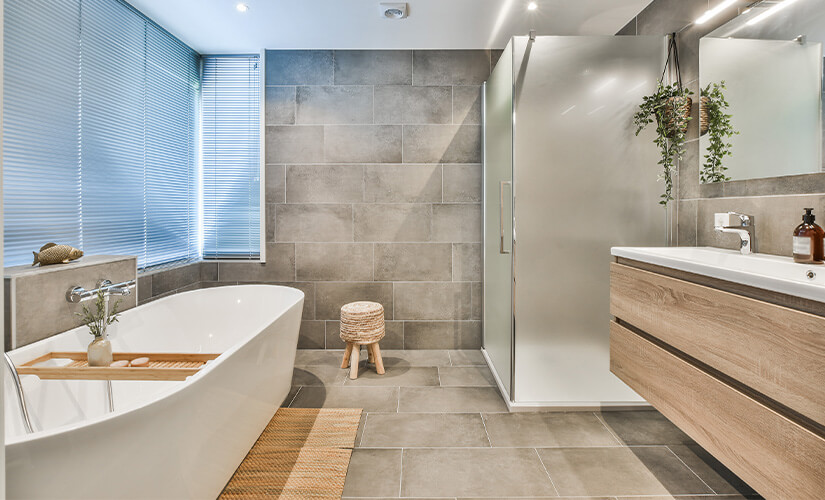 Natural wood floating vanity in modern bathroom with gray wall and floor tiles and soaker tub.