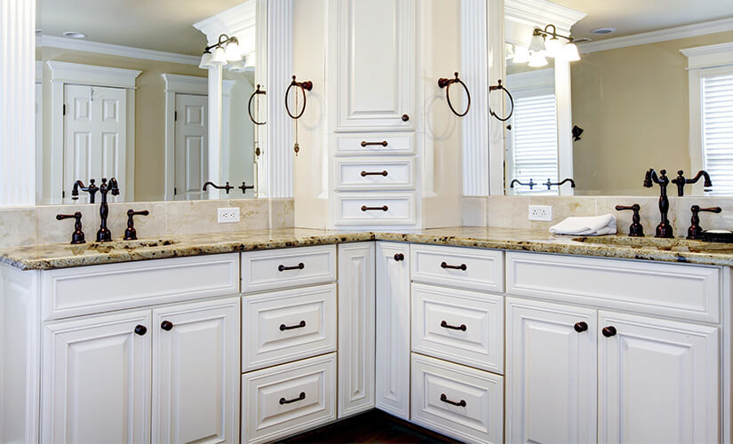 Corner bathroom vanity with white cabinets and brown granite countertops opposite window.