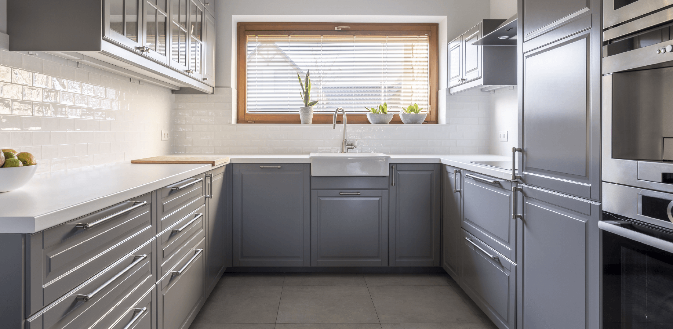 Naturally lit kitchen with gray cabinets, white countertops, farmhouse sink, and plants on the windowsill.