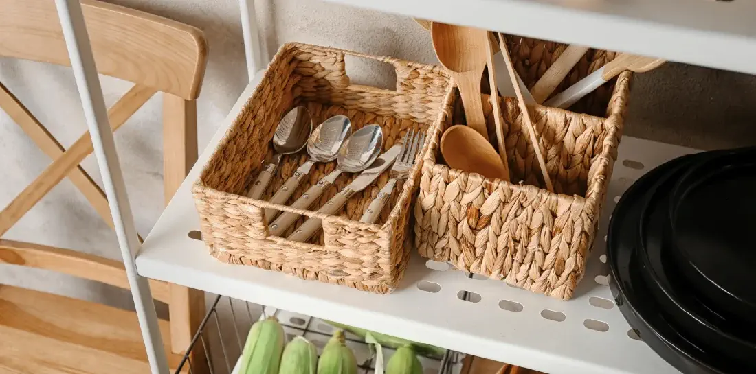 Two wicker baskets containing metal and wood utensils on a white kitchen shelf.