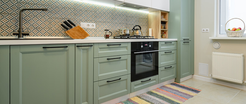 Image shows colorful kitchen with green cabinets, a colorful tile backsplash, and a colorful striped rug.