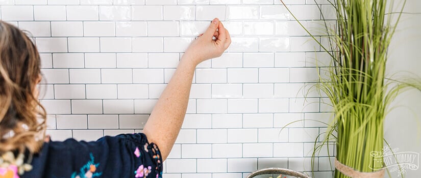 Woman applying white subway tile peel-and-stick backsplash to kitchen wall.