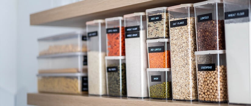Dry ingredients organized and labeled in clear plastic containers on open shelf in kitchen.