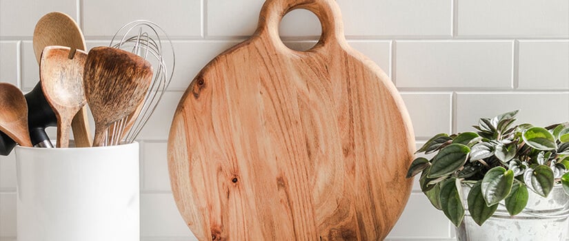 Large circular wood cutting board resting against white tiled kitchen wall between container of wooden cooking utensils and plant.
