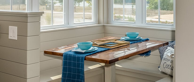 Small wood rectangular dining table with cream built-in benches below large kitchen windows.
