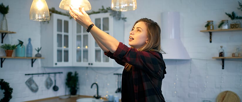 Woman updating lightbulbs in glass pendant light fixtures in kitchen.
