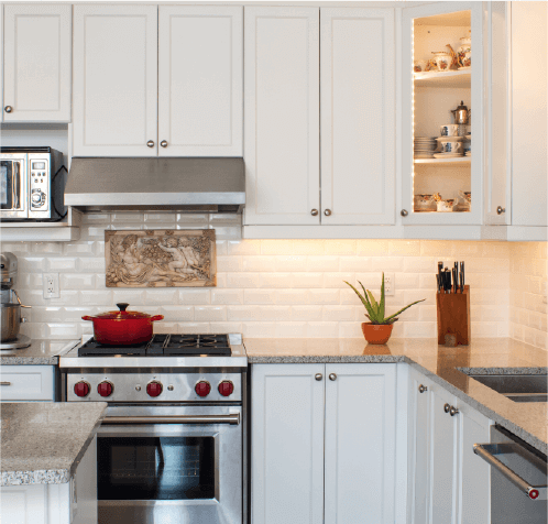 White painted cabinets in clean kitchen with stainless steel oven, and lit backsplash.