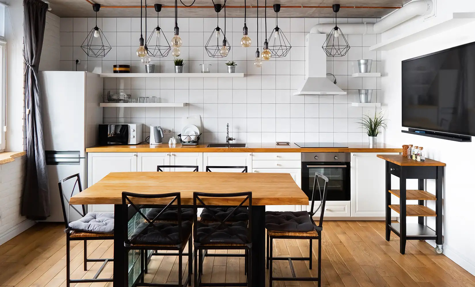 Modern industrial kitchen with wood flooring, dining table, hanging light fixtures, and a white tile backsplash.