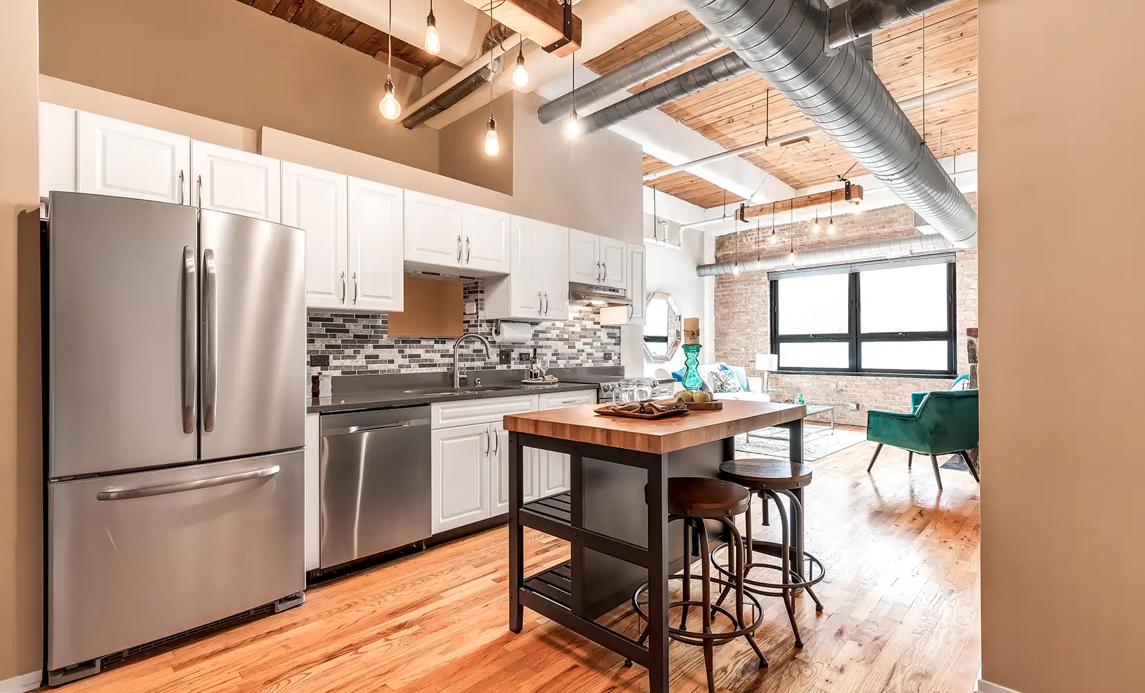 Industrial kitchen with metal ceiling beams, kitchen island, wood floors, stainless steel appliances, and hanging lights.