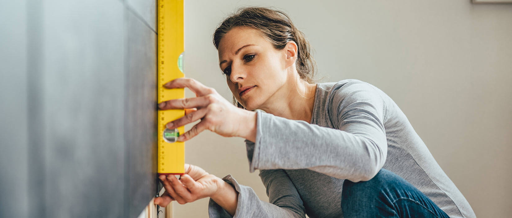 Woman in gray long sleeve shirt using yellow level to measure wall.