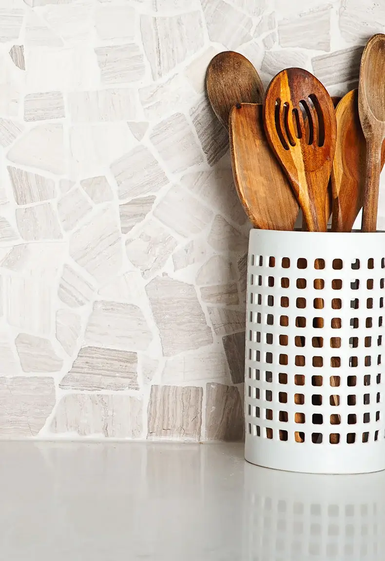 Wooden kitchen utensils in a white basket on white countertop against a white and brown-toned rock stone backsplash.