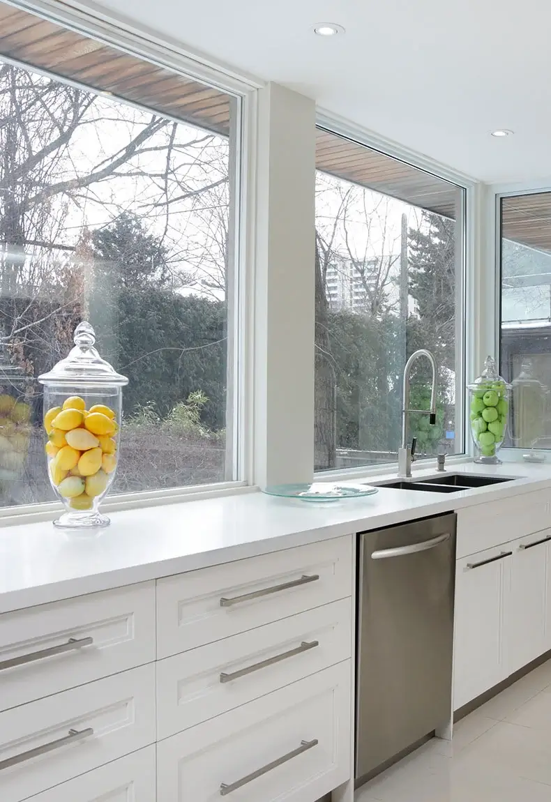 Kitchen with white cabinets, dishwasher, white countertops and large window backsplash with nature background.