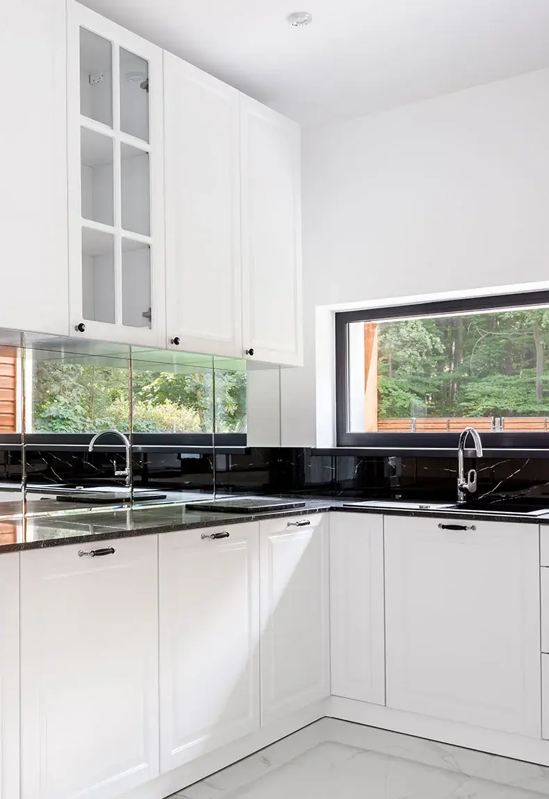 Kitchen with black countertops, white cabinets and drawers, windows with nature background, and reflective backsplash.