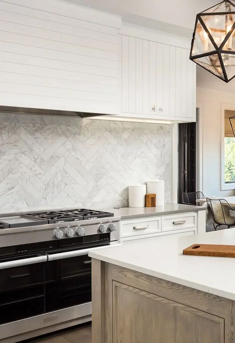 Modern kitchen interior with patterned backsplash, marble counters, kitchen island, white cabinets, and large windows.