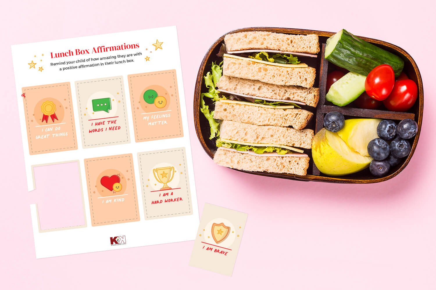 Printable lunch box affirmation cards next to a packed lunch box with sandwiches, fruits, and veggies on a pink background.