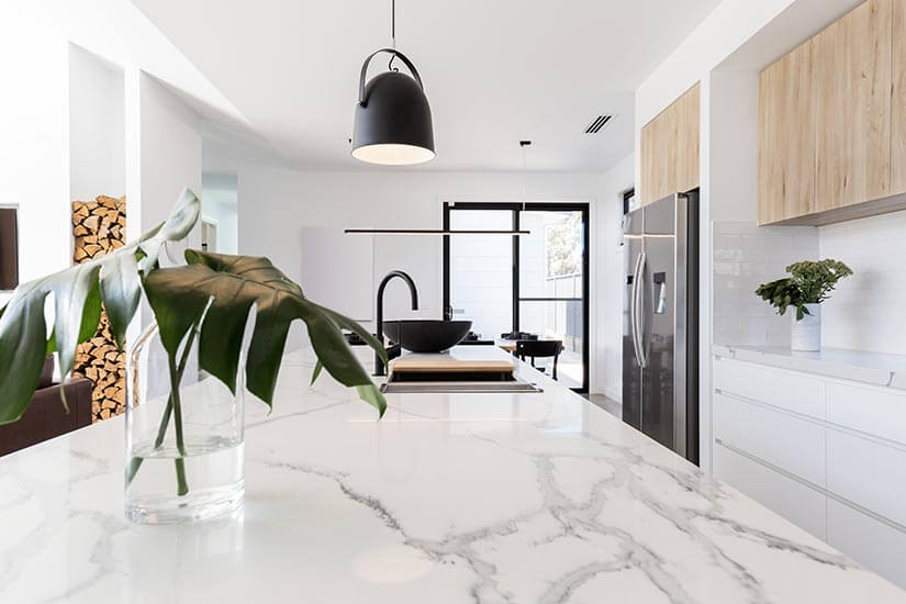 Modern kitchen with marble countertops, light wood upper cabinets, white base cabinets, and monstera leaves in glass vase on countertop.
