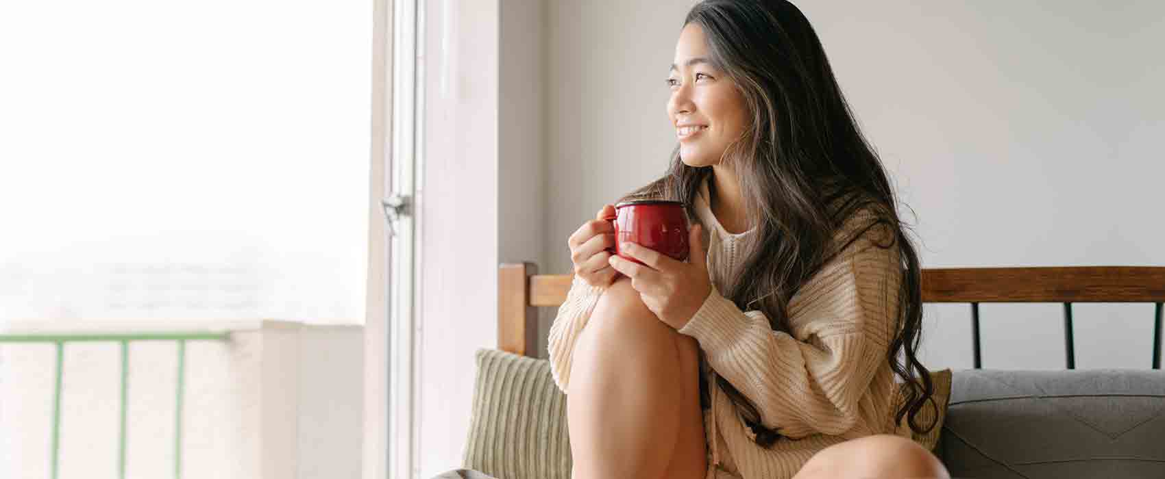 Young woman holding a mug and sitting on her bed looking out the window.