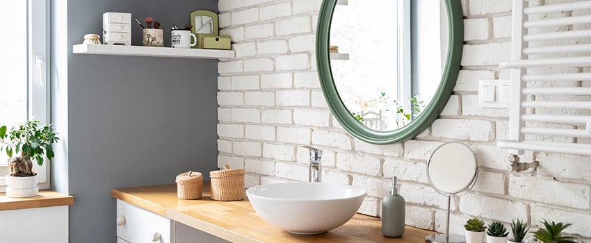 Small bathroom with white brick wall, green circle mirror, and white bowl sink on wood countertop.