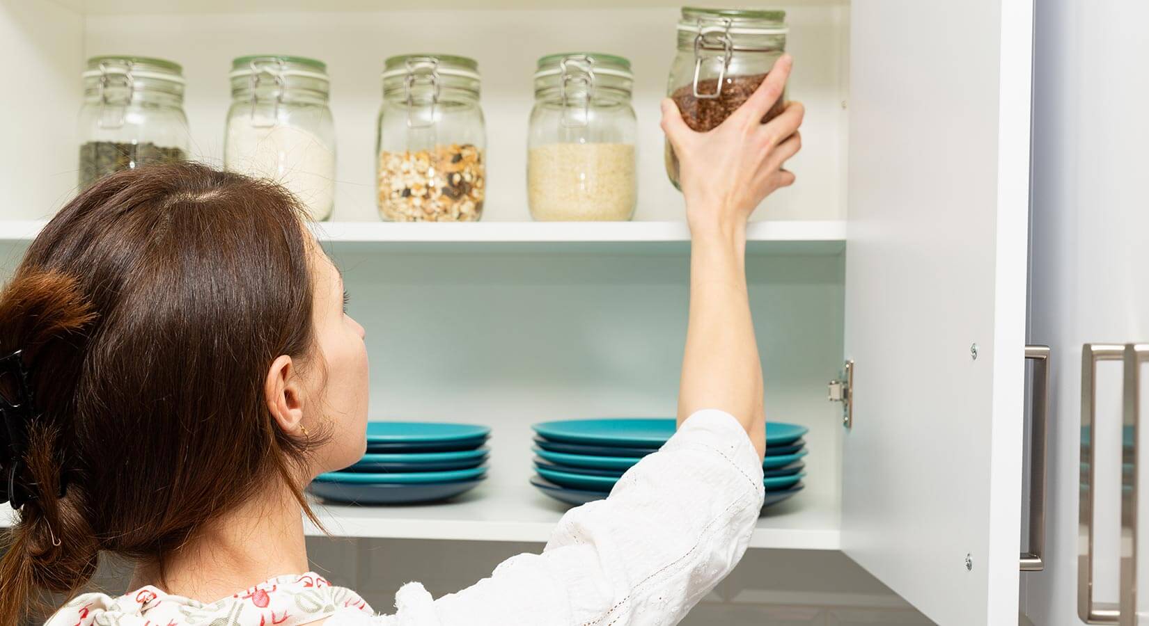 Women placing dry ingredients in glass jars inside white kitchen cabinets above blue plates.