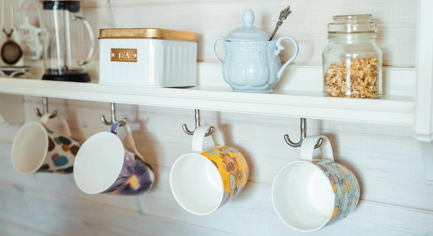 Close-up of white kitchen shelf with four colorful mugs hanging from hooks below.