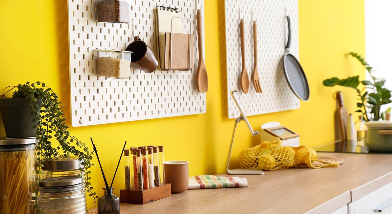Cooking utensils, pan, and mug hanging from white pegboard against bright yellow kitchen wall.