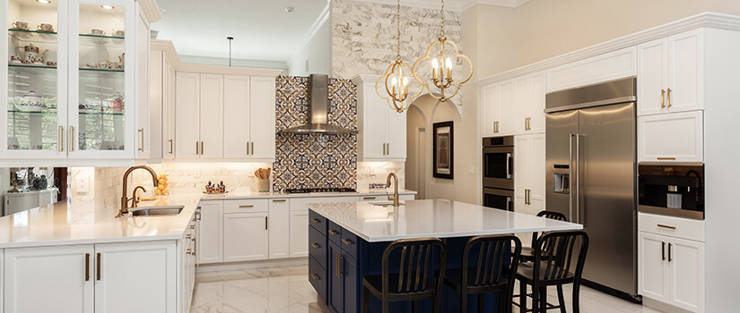 Elegant white kitchen with shaker cabinets marble floor and dark blue kitchen island in the center under golden light fixtures.