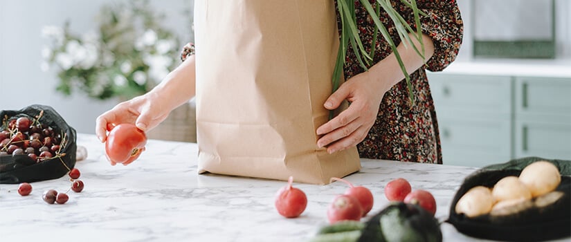 Women setting tomatoes from brown paper bag onto marble kitchen countertop.